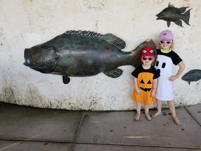 Two small kids, one with a spiderman hat and pumpkin dress, and the other with a purple hat and a ghost dress, stand in front of a large fish statue 