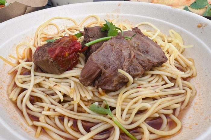 three chunks of dark brown beef shank garnished with parsley rest on top of a pile of noodles in a dark broth