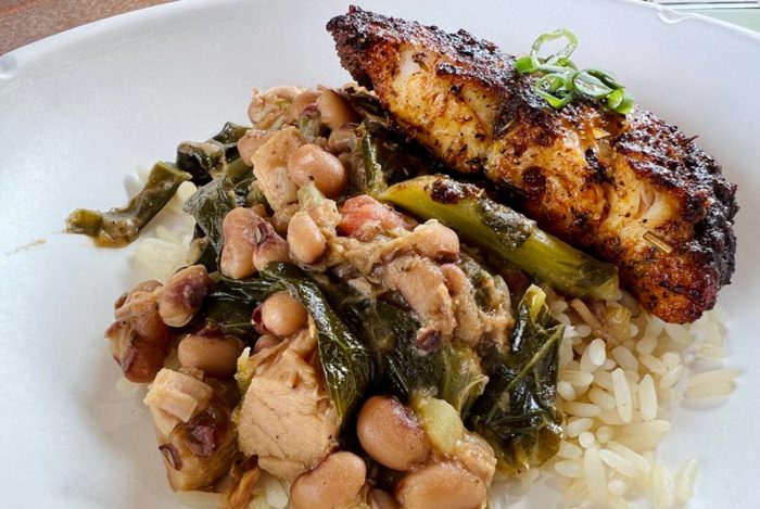 A slice of blackened catfish, slightly glistening with oil, is towards the back of the plate. It rests on a bed of rice, and forward on the rice is a serving of hoppin' john with visible beans and collards.
