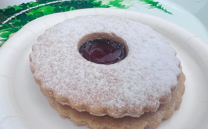 a round sandwich cookie with crinkled edges and a dusting of powdered sugar surrounding a thumbprint of deep reddish purple raspberry jam.