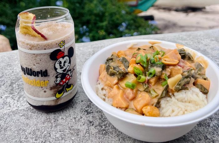 stew with peanuts, sweet potato chunks, and wilted mustard greens on a bed of rice, topped with sliced scallions. On the left a frozen brown beverage with a red straw in a glass cup with Minnie Mouse on the side