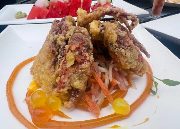 Two halves of a crispy-coated fried soft shell crab are balanced against each other, supported by a small mount of orange and white slaw-like salad underneath. A thin rope of orange sauce with occasional red flecks surrounds the whole thing, and a few bob-style yellow and orange beads are arranged at the tail of the sauce curl, where it meets the crab.