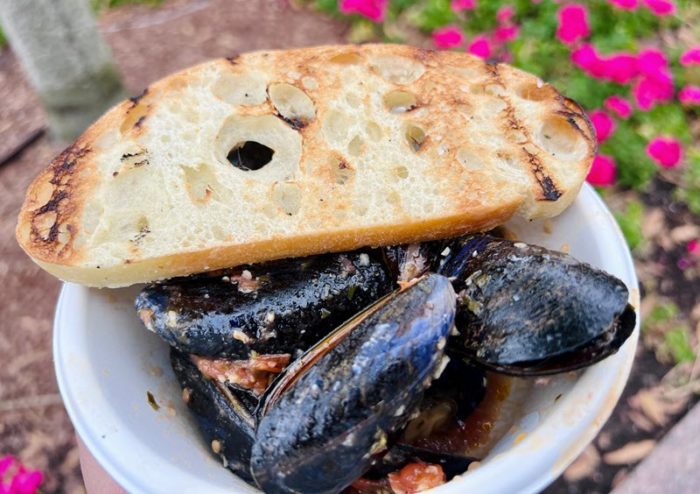 A white cardboard bowl holds several mussels with small white flecks of garlic on the shells; bits of dark red tomatoes are visible between the mussels and a piece of sliced baguette toast with grill marks lies across the top