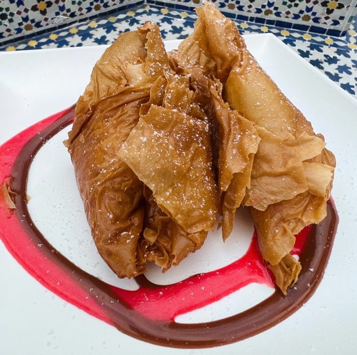 Two dark brown fried pastries lie on a plate within overlapping circles of a brown and dark pink sauce. The exterior of the pastries is fried phyllo showing ruffles and folds and looking super-crispy
