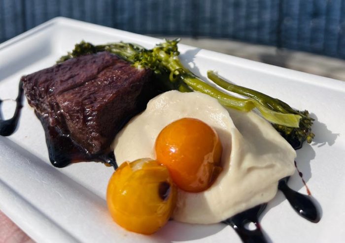 Two roasted yellow-orange baby tomatoes sit in the foreground atop a large dollop of creamy beige parsnip puree with a drizzle of balsamic peeking out from underneath. To the rear, two dark brown short ribs lie on top of more balsamic reduction with two spears of broccolini laid alongside everything.