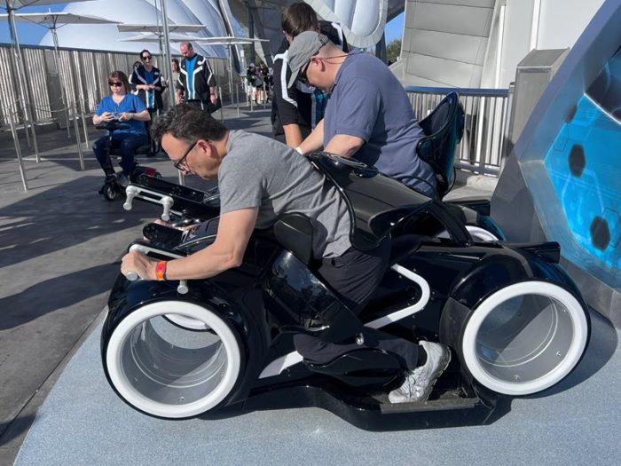 a man sits on the test vehicle for the tron ride. he is leaning forward with his hands on the handlebars as though riding a bike. His body is about 30 degrees from horizontal, and a restraint curves around his back, holding him into the seat.