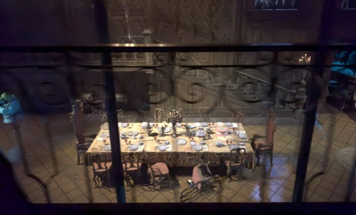 seen from above, a dining room displays a formal place setting with several chairs tipped over. To the far left, dancing ghosts can be seen.