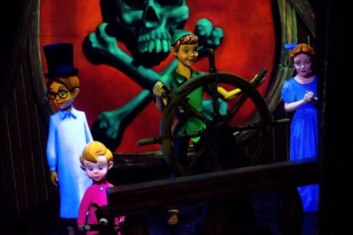 Peter Pan stands at the helm while the Darling children look on. Behind him is a large red flag with a green skull and crossbones.